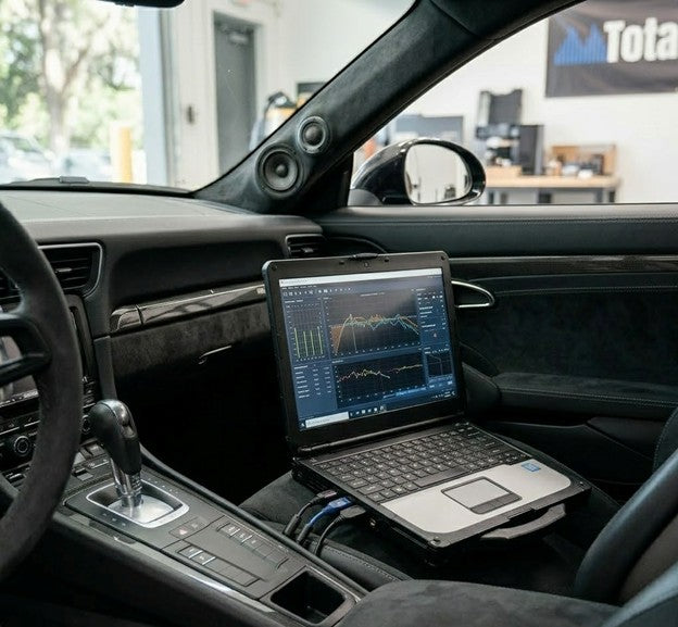 A laptop displaying professional car audio DSP tuning software sits on the driver's seat of a high-end vehicle, with visible acoustic measurements on the screen and a Total Tuning banner in the background.